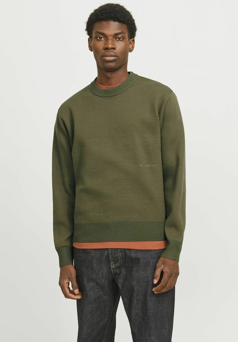 Suéter verde de punto con un cuello redondo de canalé y puños. Lleva un camisa de color óxido debajo, combinado con jeans de mezclilla oscuros.