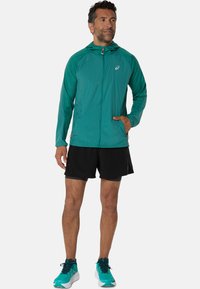 Veste zippée légère turquoise avec capuche, dotée d'une fermeture éclair frontale et de poches latérales, associée à un short noir et des chaussures de course turquoise.
