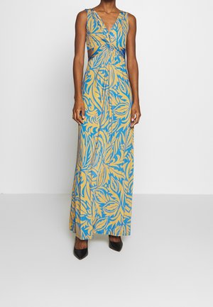 Kobieta ubrana w niebiesko-beżową sukienkę bez rękawów sięgającą podłogi z motywem liści i marszczeniem na wysokości piersi, w połączeniu z czarnymi szpilkami.