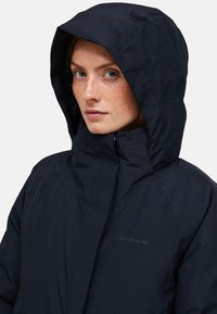 Vrouw met een lichte huid en sproeten, gekleed in een donkere marineblauwe waterdichte jacket met capuchon, tot aan de hals dichtgeritst, tegen een lichte achtergrond.