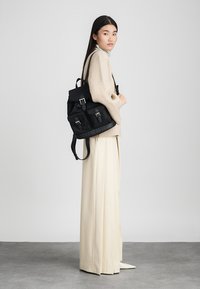 Zwarte canvas rugzak met metalen details, twee voorzakken en verstelbare bandjes. Draag het met een beige sweater en brede crèmekleurige broek.