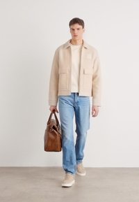 Chaqueta de forro polar beige con dos bolsillos delanteros, suéter beige, jeans azul claro y zapatillas de ante. Bolsa de cuero marrón en la mano.
