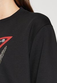 Sudadera negra con cuello redondo acanalado. Presenta un diseño de logo rojo con adornos de cuentas en un patrón en zigzag en la parte frontal.