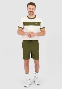 Männeroutfit mit einem cremefarbenen T-Shirt mit grünen Streifen und Logo, kombiniert mit olivgrünen Shorts. Sneaker und Socken runden den Look ab.