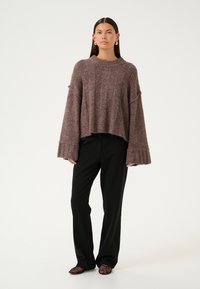 Chunky brown knit sweater with wide sleeves, raw hem, worn over black trousers, paired with textured black and red shoes, against a plain background.