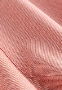 Tekstil i et blødt, vævet mønster med en koralrosa nuance; viser subtil tekstur og diagonale folder, omkranset af en fin stikningsdetalje.