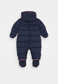 Marineblauwe puffy sneeuwoverall met een capuchon. Beschikt over een gevoerd ontwerp, elastische manchetten en rode afwerking bij de polsen en enkels. Ritssluiting op de rug.