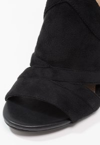 Chaussure noire en daim à bout ouvert avec des brides superposées et une semelle à texture lisse.