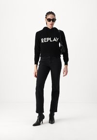 Haut à capuche cropped côtelé noir avec le logo "REPLAY", associé à un jean noir taille haute et des bottines à talons noires. Les accessoires comprennent des lunettes de soleil noires et un petit sac noir.