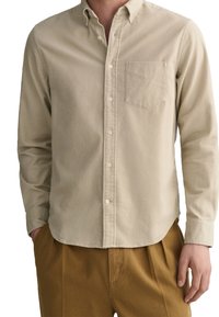 Beige knappad skjorta med långa ärmar, bröstficka och en mjuk textur; parat med tanfärgade byxor, vilket visar en avslappnad stil.