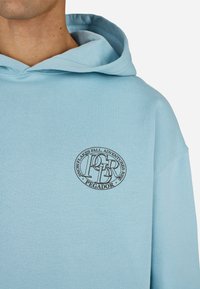 Ανοιχτό μπλε φούτερ από μαλακό ύφασμα. Διαθέτει κυκλικό μαύρο λογότυπο στο στήθος με κείμενο. Κουκούλα με χαλαρή εφαρμογή.