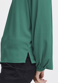 Blusa verde, leggera, a maniche lunghe con polsini arricciati e spacchi laterali. Texture liscia; presenta una silhouette rilassata.