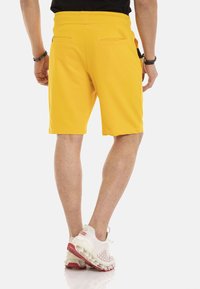 Gele sportieve shorts met een gladde textuur, elastische tailleband en twee achterzakken, gecombineerd met witte en rode hardloopschoenen.