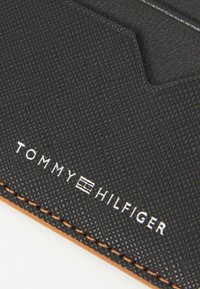Portofel din piele texturată neagră cu margine bej, având logo-ul embosat "TOMMY HILFIGER" în alb și un design distinctiv cu decupaje geometrice.