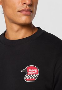 Close-up of a person wearing a black shirt with a red and black checkered helmet design and "Replay Helmet" text on the chest.