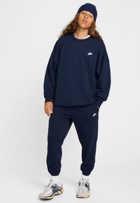 Tumši zils Nike sporta krekls un bikses ar baltu logo uz krūtīm un augšstilba, kopā ar gaišiem sporta apaviem un atbilstošu cepuri.