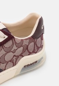 Gros plan d'une basket bordeaux à motif avec doublure beige, languette de talon brun foncé et semelle translucide avec le logo Coach.