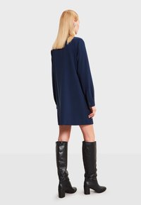 Žena so rovnými blond vlasmi, oblečená v dlhých modrých šatách s dlhými rukávmi a čiernych čižmách na opätku po kolená, stojaca so chrbtom k nám.