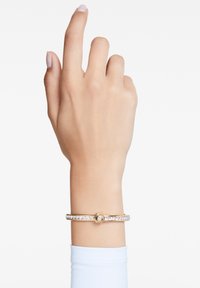 Pulsera en tono dorado con un centro hexagonal, que presenta una línea de cristales claros a lo largo de la banda. Textura suave, diseño minimalista.