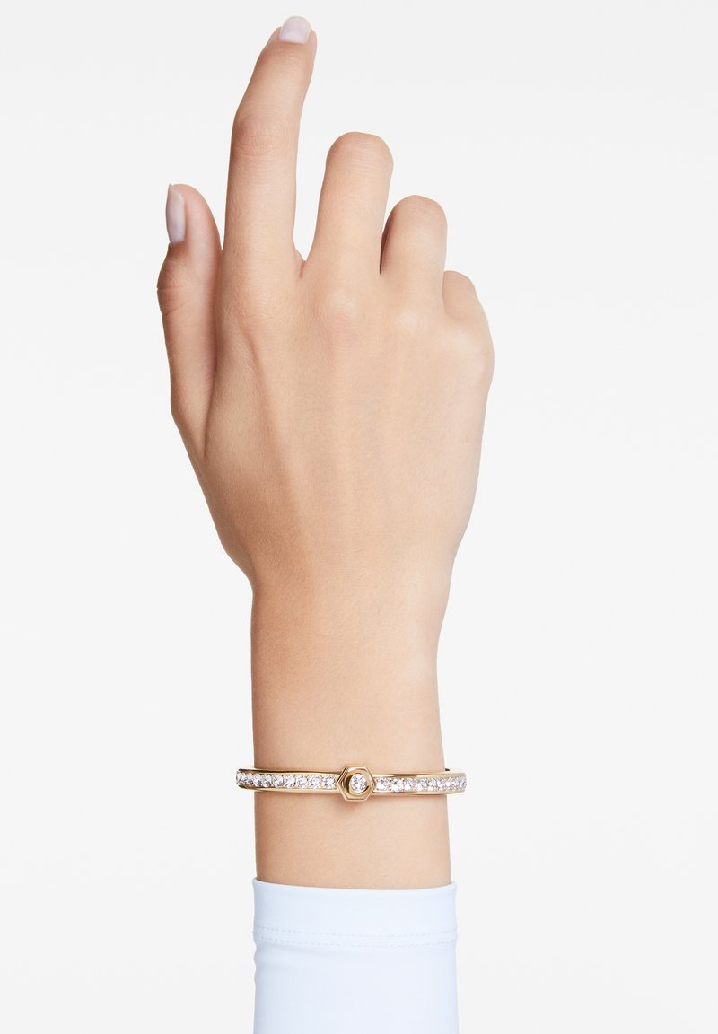 Pulsera en tono dorado con un centro hexagonal, que presenta una línea de cristales claros a lo largo de la banda. Textura suave, diseño minimalista.