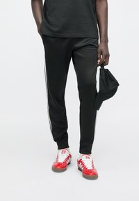 Čierne úzke teplákové nohavice s bielymi pruhmi na bokoch, doplnené červenými športovými topánkami a čiernou čiapkou drženou v ruke. Hladký textilný povrch.