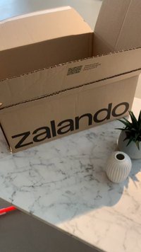 Zalando-kartonboksen har et dristig svart logo, plassert på en marmoroverflate ved siden av en teksturert hvit vase og en liten potteplante.