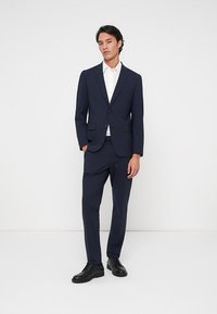 Άνδρας που στέκεται φορώντας ένα ναυτικό προσαρμοσμένο κοστούμι, λευκό πουκάμισο και μαύρες δερμάτινες γόβες σε έναν σκέτο λευκό φόντο.