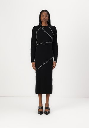 Black knit dress with long sleeves, featuring white wavy stitching. Below the knee length, paired with black high-heeled shoes with buckles.
