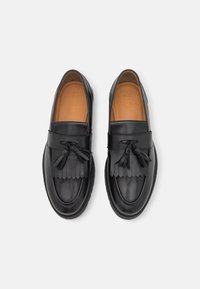 Mocassins en cuir noir avec des détails à franges et des pompons. Texture lisse, bout rond et semelle intérieure beige contrastante. Design classique pour un port polyvalent.