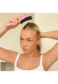 Czarna szczotka z włosiem i różową rączką trzymana nad głową osoby. Włosy są przedzielone i zaczesane do tyłu, na tle neutralnej ściany.