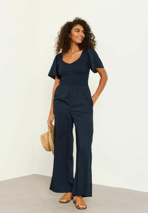 Frau mit lockigem Haar, die einen marineblauen Overall trägt, einen Strohhut hält und geflochtene Sandalen trägt, vor einem schlichten Hintergrund stehend.