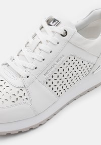 Baskets blanches Michael Kors avec panneaux latéraux perforés, lacets blancs et détail du nom de la marque en argent sur la tige en cuir.