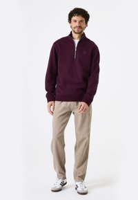 Bordeaux zip-up sweatshirt met ribgebreide manchetten, gecombineerd met lichtbeige corduroy broek en zwart-witte sneakers. Geen zichtbare patronen.