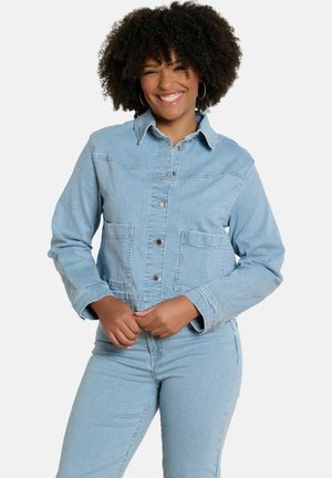 Uśmiechnięta kobieta o kręconych włosach, nosząca kolczyki-koła, jasnoniebieską, w paski marynarkę i pasujące spodnie z wysokim stanem, stojąca na białym tle.