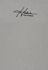 Grå bomulls t-shirt med en svart broderad "Hollister California"-logotyp på övre vänstra sidan. Slät textur med en enkel design.
