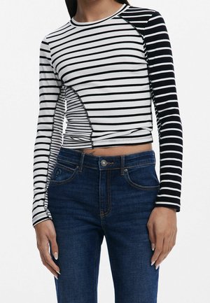 Mujer que lleva una camiseta corta de manga larga a rayas negras y blancas con patrones asimétricos y vaqueros de talle alto de color azul oscuro.