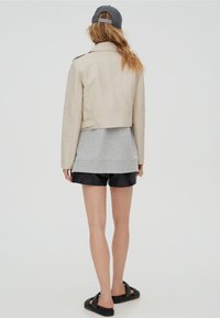 Beige Kunstleder cropped Jacke mit Schulterklappen, getragen über einem grauen Sweatshirt, schwarzen Shorts und schwarzen Sandalen. Neutraler Hintergrund.