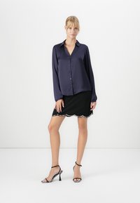 Woman standing wearing navy buttoned blouse, black skirt with scalloped hem, and black high-heeled sandals with zebra pattern heels.