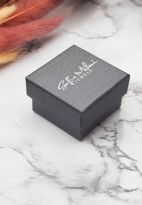 Piccola scatola di gioielli nera con texture, con il logo "Sofia Milani JEWELS" su una superficie di marmo bianco, con erba pampas asciutta di colore rosa e beige nelle vicinanze.