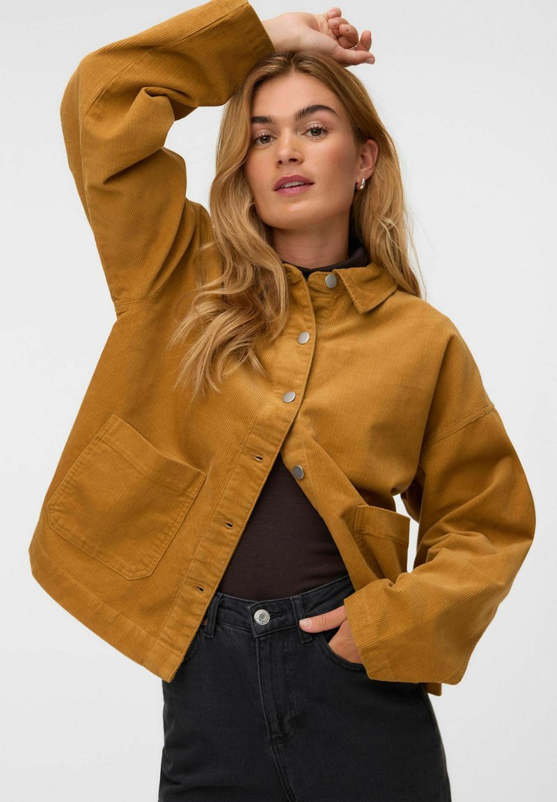 Femme portant une veste en velours côtelé moutarde avec boutons-pression, pantalon noir et haut noir, posant avec un bras levé au-dessus de sa tête.
