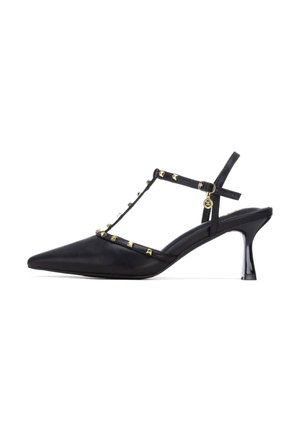 Zapato de tacón negro de cuero con puntera puntiaguda, diseño de tira en T, tachuelas piramidales doradas y tacón delgado y brillante.