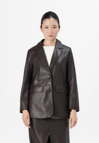 Vrouw in een donkerbruin leren blazer over een witte top en een bijpassende leren rok, staand tegen een effen witte achtergrond.