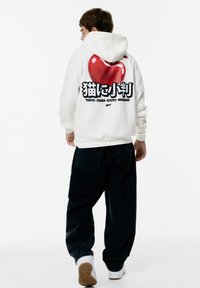 Felpa bianca con cappuccio e grafica di una mela rossa lucida sul retro. Il testo nero include caratteri giapponesi e nomi di città. Pantaloni neri e sneakers bianche.