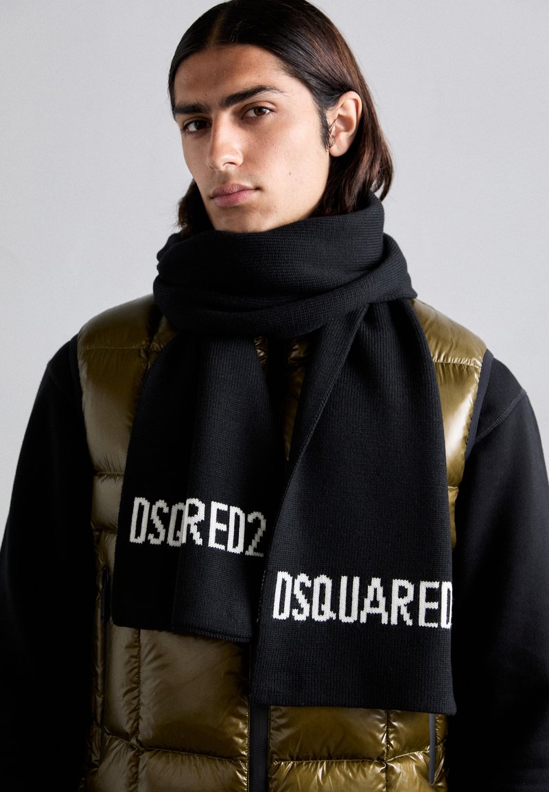 Scaldino nero a maglia con logo "DSQRED2" in bianco, indossato allentato attorno al collo, abbinato a un gilet imbottito lucido di colore verde oliva.