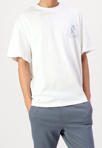 White cotton t-shirt with short sleeves, round neckline, and blue graphic detail on the left chest. Paired with light blue trousers.