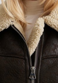 Brun jakke i imiteret læder med en fluffy beige shearling-krave og lynlås lukning, der har synlige syninger og en tekstureret overflade.