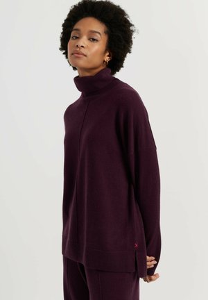 Vrouw met kort krullend haar, die een losse donkerpaarse coltrui en bijpassende broek draagt tegen een effen achtergrond.