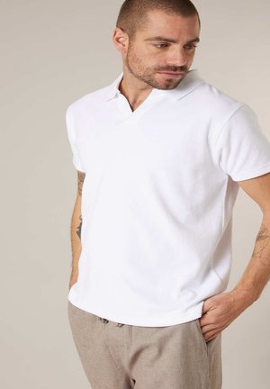 Homme aux cheveux courts et barbe portant un polo blanc à manches courtes et un pantalon beige, debout les mains dans les poches, regardant vers le bas à droite.