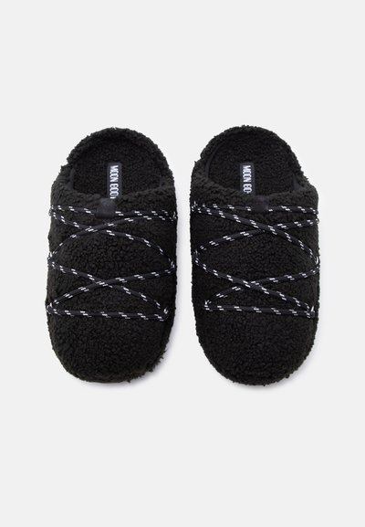 Chaussons en polaire noire à surface texturée, avec des sangles élastiques blanches croisées et un design arrondi. L'étiquette indique "MOON GOOD."