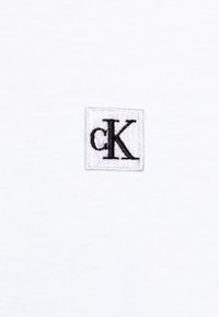 Vit bomullstyg med en fyrkantig broderad logotyp som har små bokstäver "c" och "K" i svart. Enkel design, mjuk textur.